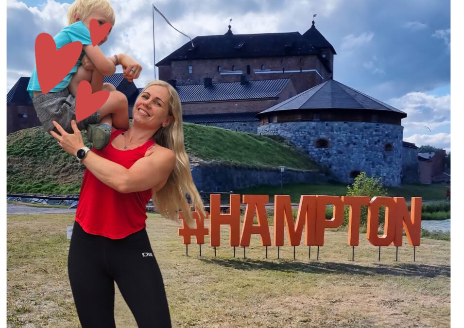 Kristiina Lindroos lapsen kanssa Hämeen linnan maiseman edessä. Taustalla kyltti, jossa lukee #HÄMPTON